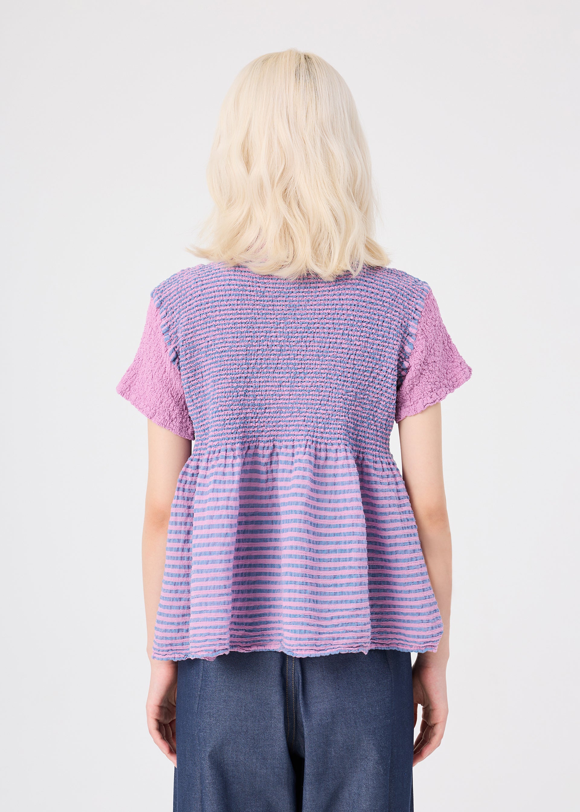 Zoomed view A person with shoulder-length blonde hair, seen from behind, wears the STRIPE CAULIFLOWER cotton short-sleeved top by me ISSEY MIYAKE—featuring purple and pink stripes and textured fabric—paired with blue pants against a plain light background.