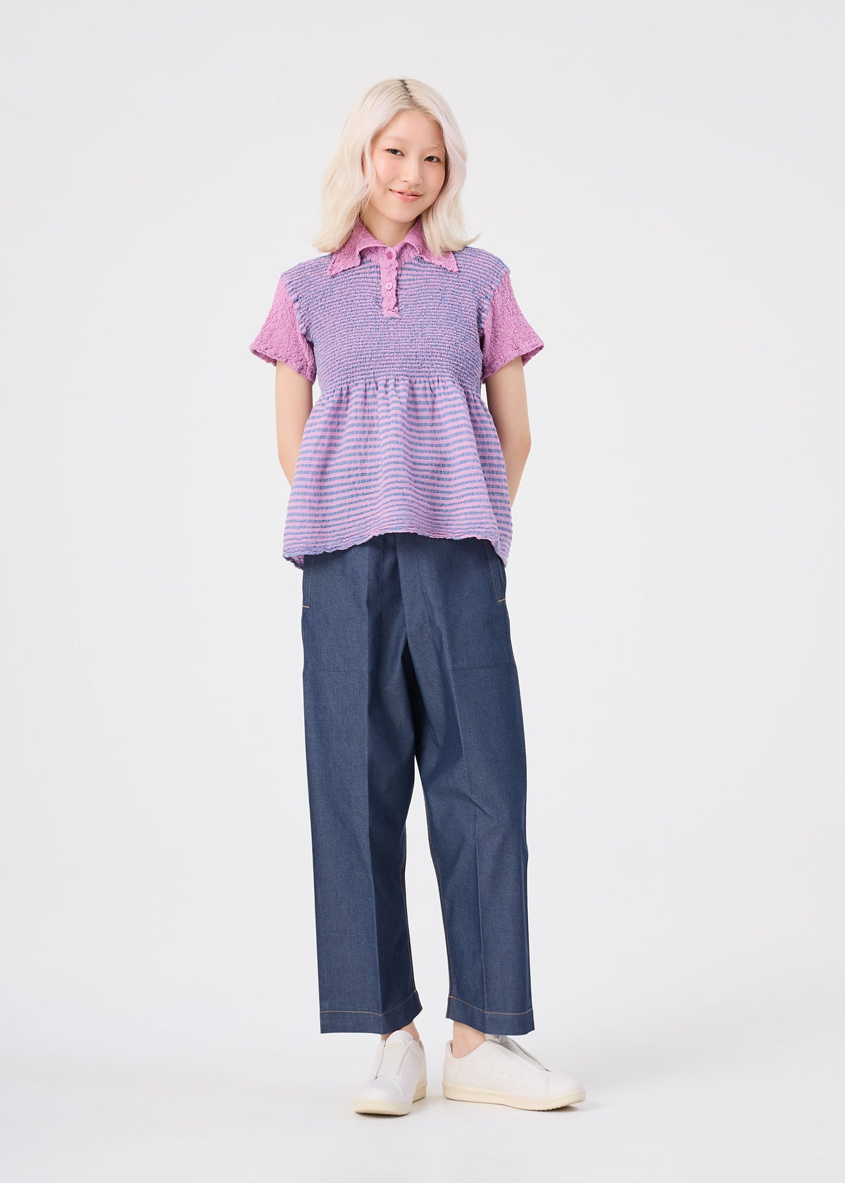 A person with light blonde hair, dressed in the me ISSEY MIYAKE STRIPE CAULIFLOWER cotton short-sleeved purple top, dark wide-leg pants, and white slip-on shoes stands against a plain white background, smiling with hands behind their back. view 7 of 7
