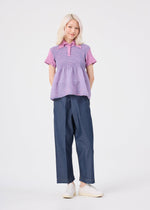 A person with light blonde hair, dressed in the me ISSEY MIYAKE STRIPE CAULIFLOWER cotton short-sleeved purple top, dark wide-leg pants, and white slip-on shoes stands against a plain white background, smiling with hands behind their back.
