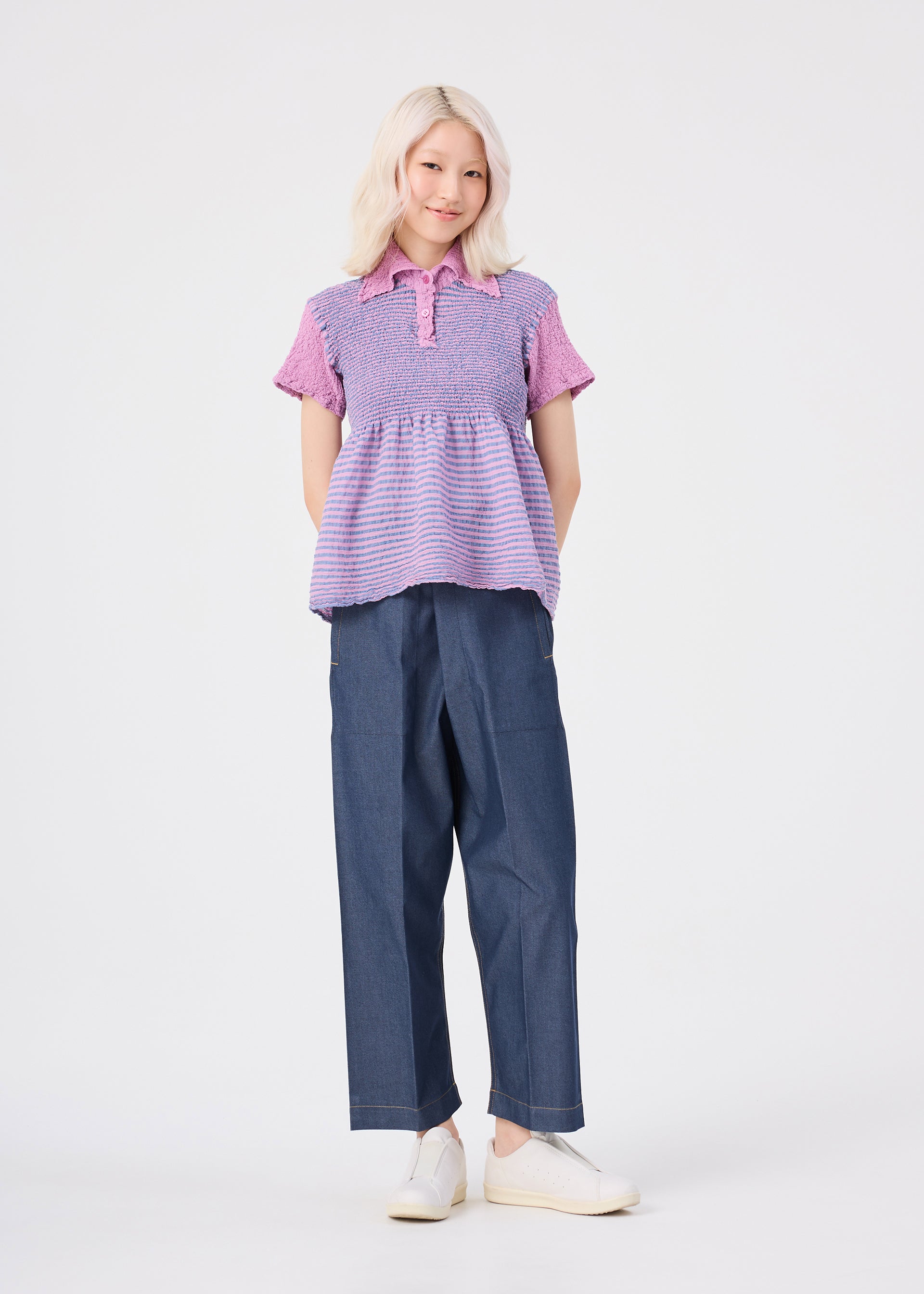 Zoomed view A person with light blonde hair, dressed in the me ISSEY MIYAKE STRIPE CAULIFLOWER cotton short-sleeved purple top, dark wide-leg pants, and white slip-on shoes stands against a plain white background, smiling with hands behind their back.