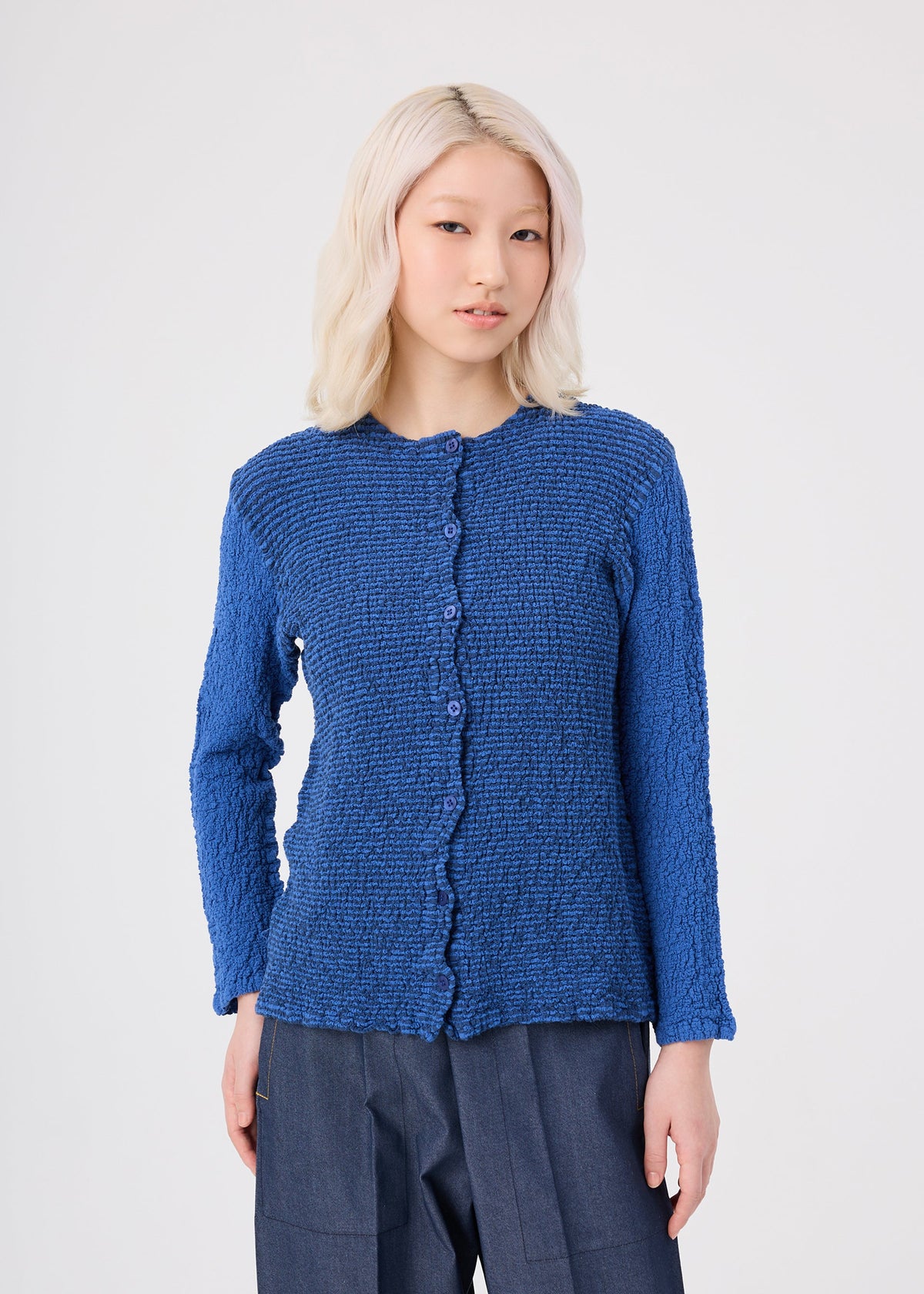 A person with light blonde hair wears the STRIPE CAULIFLOWER textured, long-sleeve blue button-up top by me ISSEY MIYAKE, made from striped fabric and paired with dark blue pants, standing against a light gray background. view 5 of 7