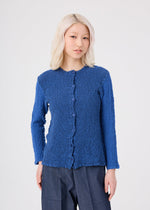 A person with light blonde hair wears the STRIPE CAULIFLOWER textured, long-sleeve blue button-up top by me ISSEY MIYAKE, made from striped fabric and paired with dark blue pants, standing against a light gray background.