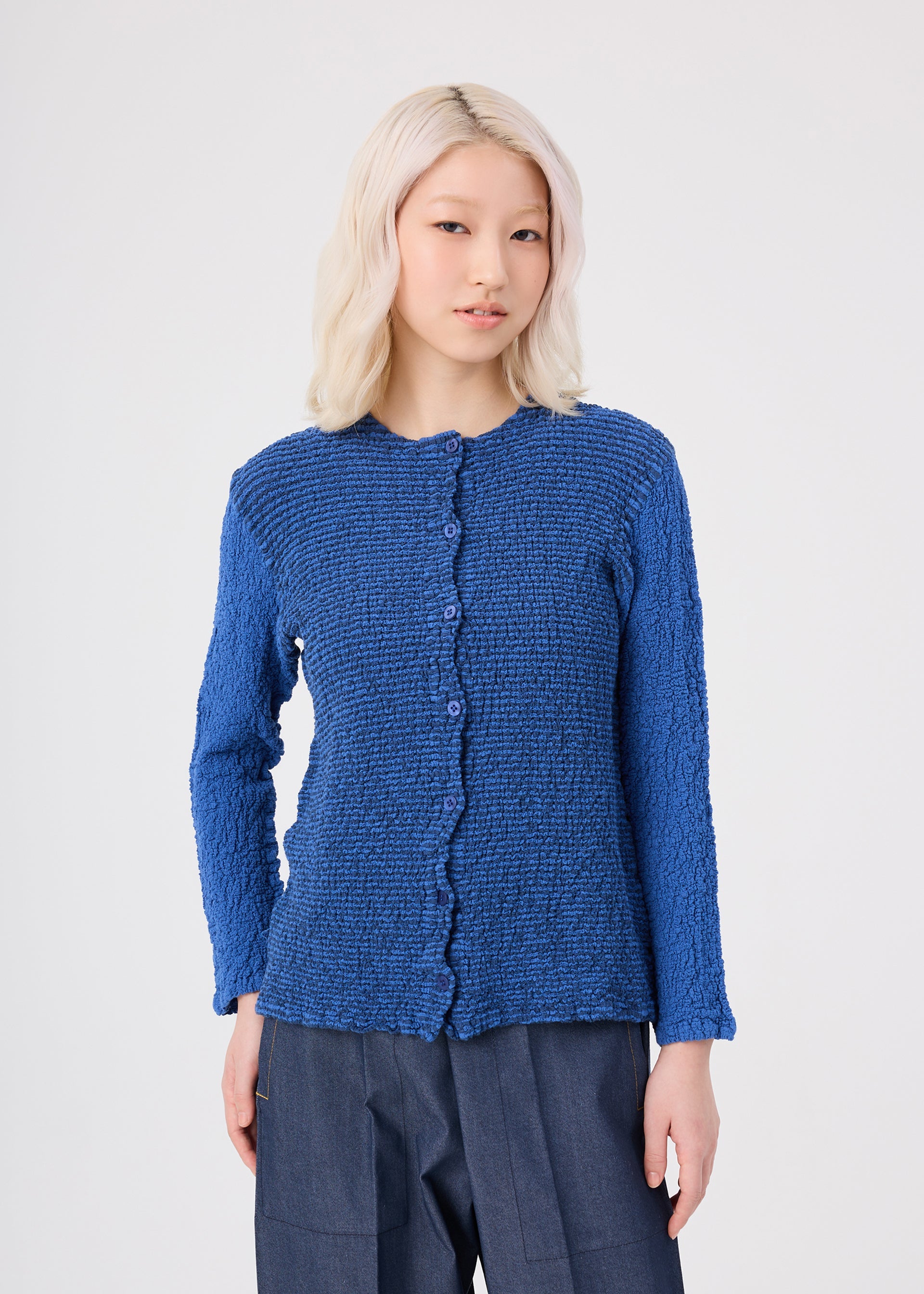 Zoomed view A person with light blonde hair wears the STRIPE CAULIFLOWER textured, long-sleeve blue button-up top by me ISSEY MIYAKE, made from striped fabric and paired with dark blue pants, standing against a light gray background.