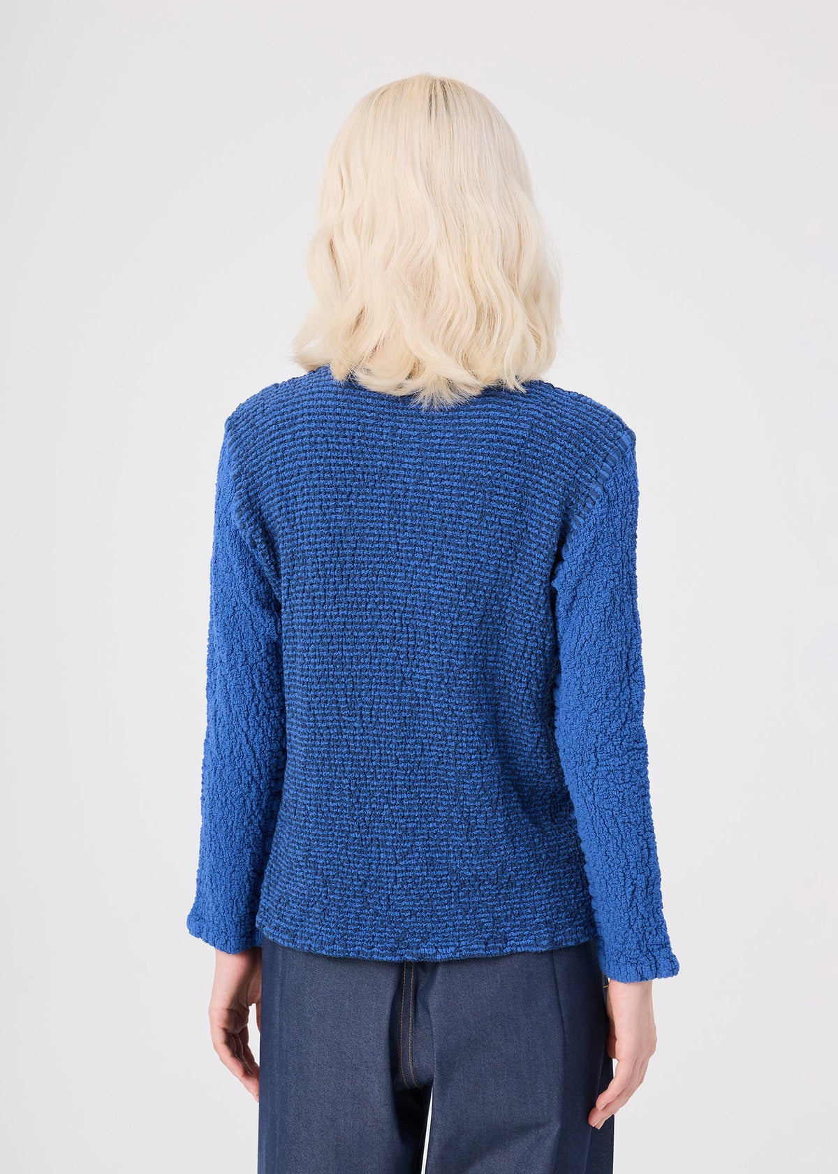 A person with wavy, shoulder-length blond hair is seen from behind, wearing the STRIPE CAULIFLOWER textured blue cotton cardigan by me ISSEY MIYAKE and dark pants, standing against a plain white background. view 6 of 7