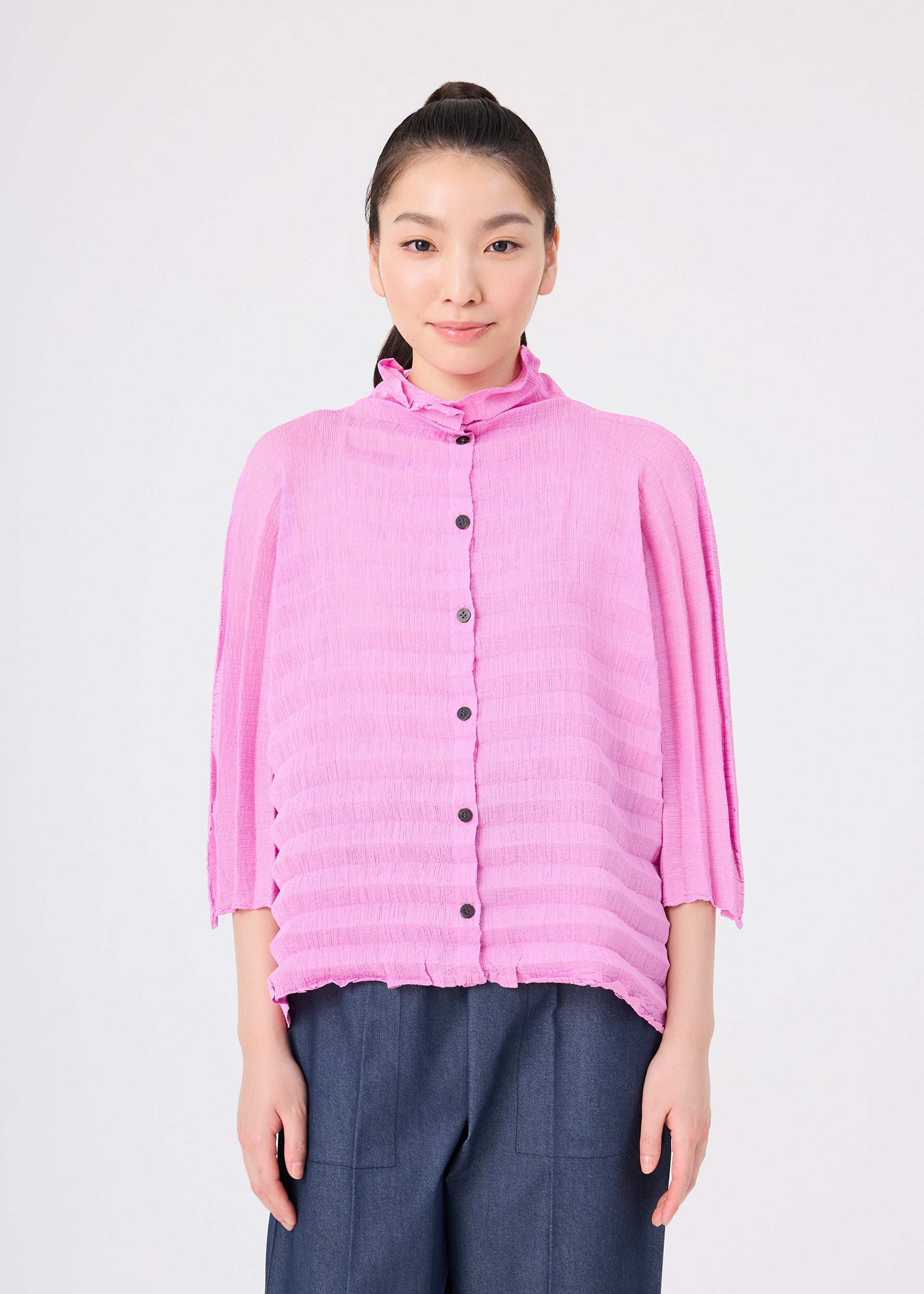 Zoomed view A person stands facing forward against a plain light background wearing the ACCORDION SEE-THROUGH CREPE top by me ISSEY MIYAKE—a textured, garment-dyed pink button-up with three-quarter sleeves—paired with blue wide-leg pants.