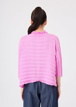 A person with long dark hair in a ponytail, facing away from the camera against a plain light background, wears the ACCORDION SEE-THROUGH CREPE top by me ISSEY MIYAKE—a textured, bright pink garment-dyed piece featuring three-quarter sleeves—paired with loose blue jeans.