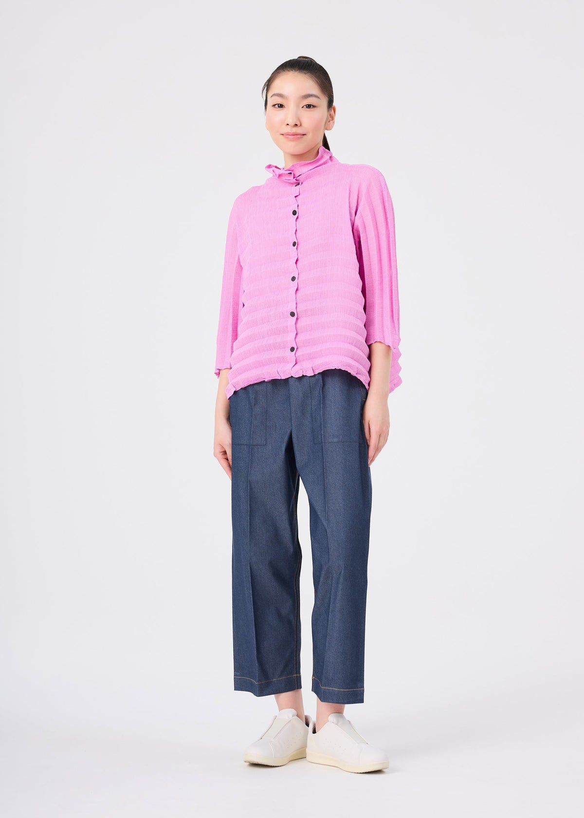 A person stands against a white background, wearing the bright pink ACCORDION SEE-THROUGH CREPE long-sleeve top by me ISSEY MIYAKE, styled with loose blue pants and white shoes. The individual looks at the camera and smiles slightly. view 7 of 7