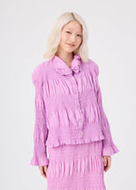 A woman with platinum blonde hair, wearing the me ISSEY MIYAKE SHRINK STRIPE light purple, textured long-sleeve cotton-blend cardigan and matching skirt, stands against a plain white background, smiling softly.