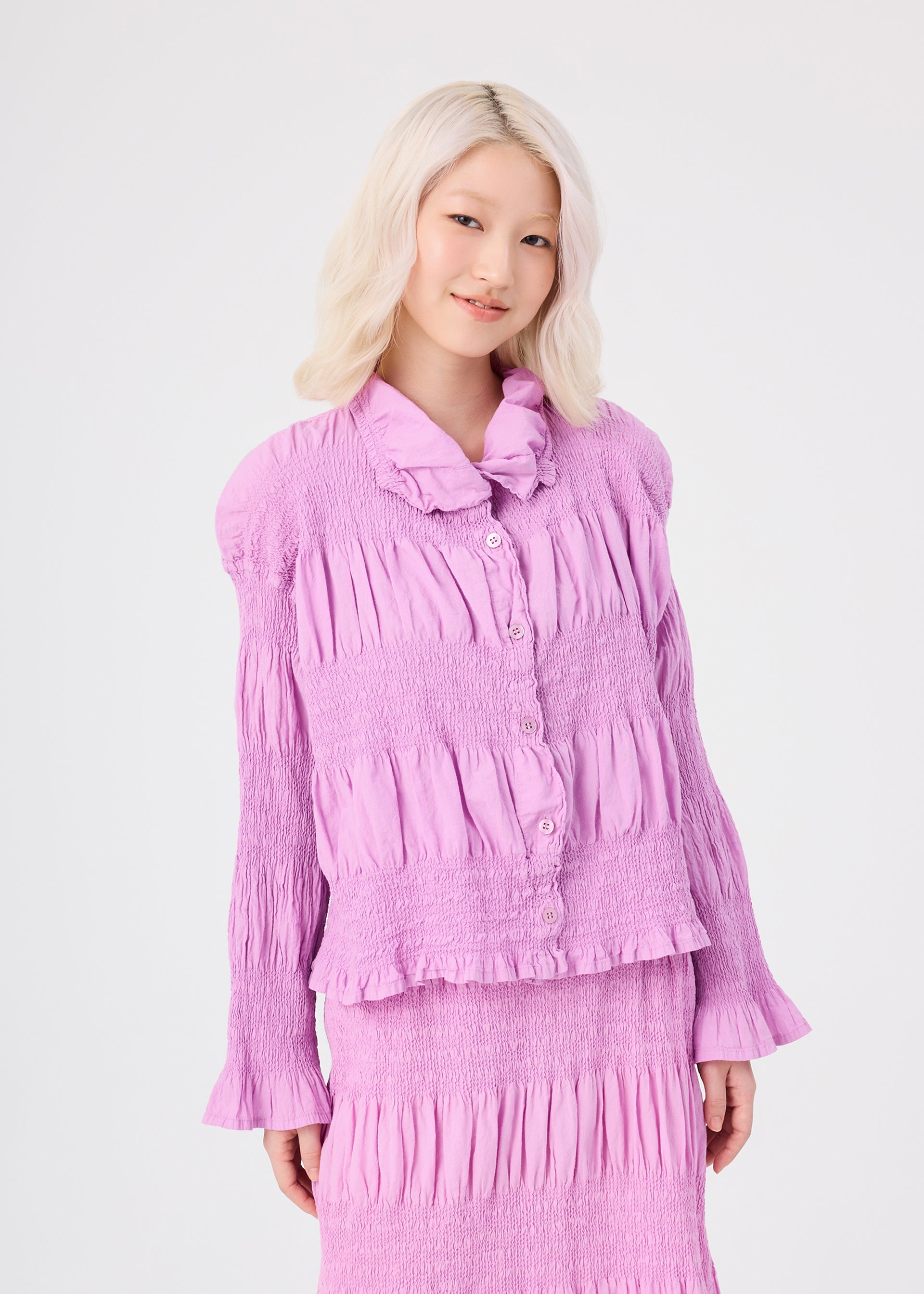 Zoomed view A woman with platinum blonde hair, wearing the me ISSEY MIYAKE SHRINK STRIPE light purple, textured long-sleeve cotton-blend cardigan and matching skirt, stands against a plain white background, smiling softly.