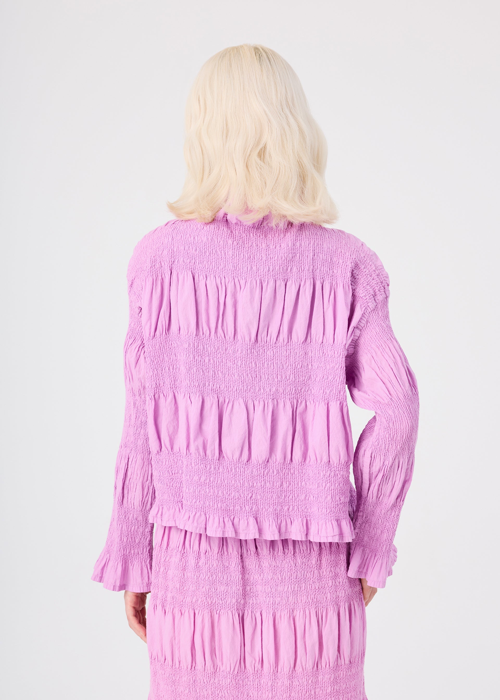 Zoomed view A person with wavy platinum blonde hair, seen from behind, wears the SHRINK STRIPE textured light purple dress with long sleeves by me ISSEY MIYAKE, paired with a soft cotton blend cardigan draped gently over their shoulders.