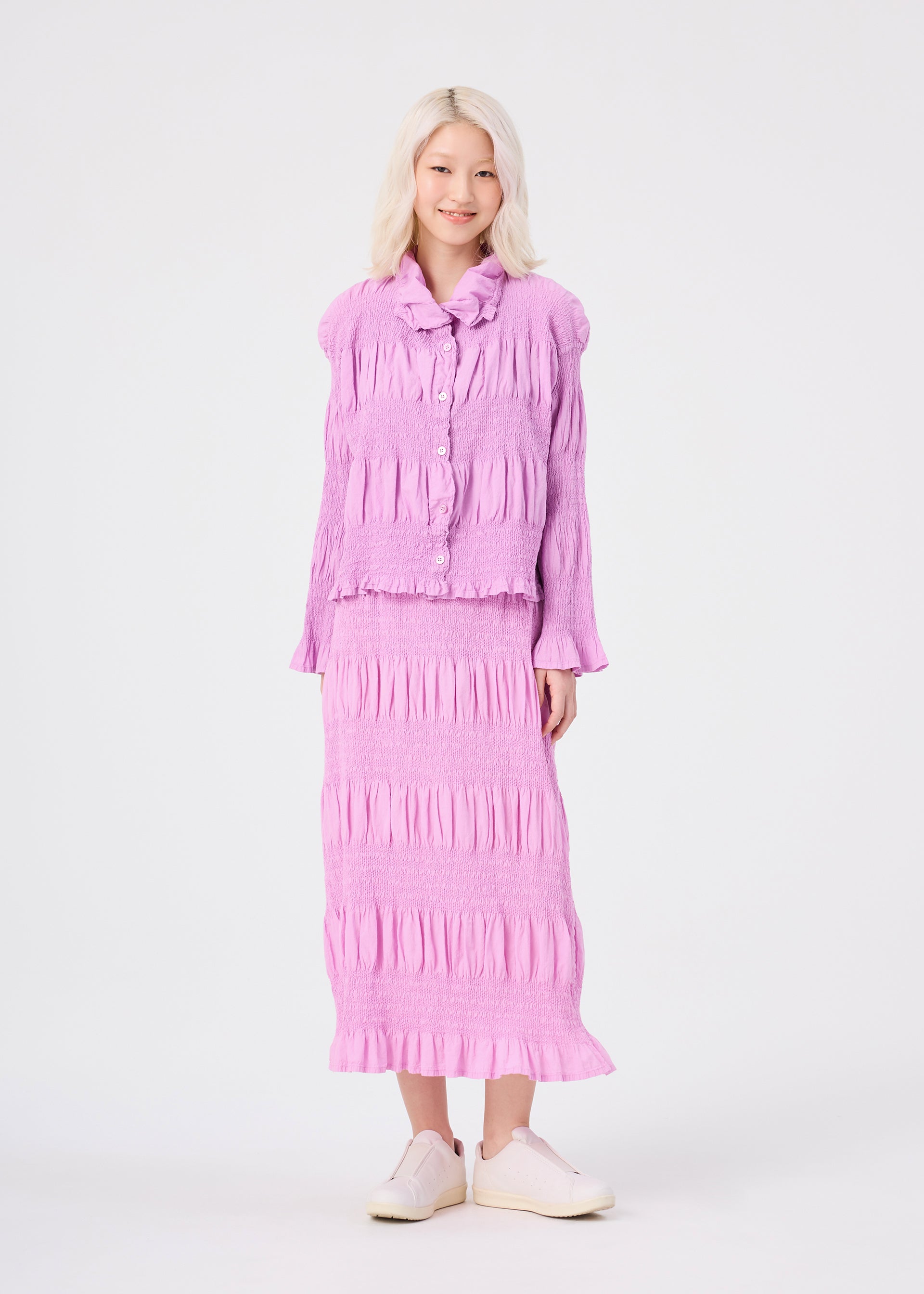 Zoomed view A person with light blonde hair models the SHRINK STRIPE textured light purple button-up top and matching long skirt with ruffled details by me ISSEY MIYAKE—both featuring a subtle product-dyeing technique—paired with white slip-on shoes, against a plain white background.