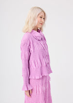 A woman with light blonde hair stands sideways against a plain white background, wearing a textured, long-sleeved lavender blouse and matching skirt, complemented by the me ISSEY MIYAKE SHRINK STRIPE cardigan, distinguished by its striped pattern and unique product-dyeing technique.