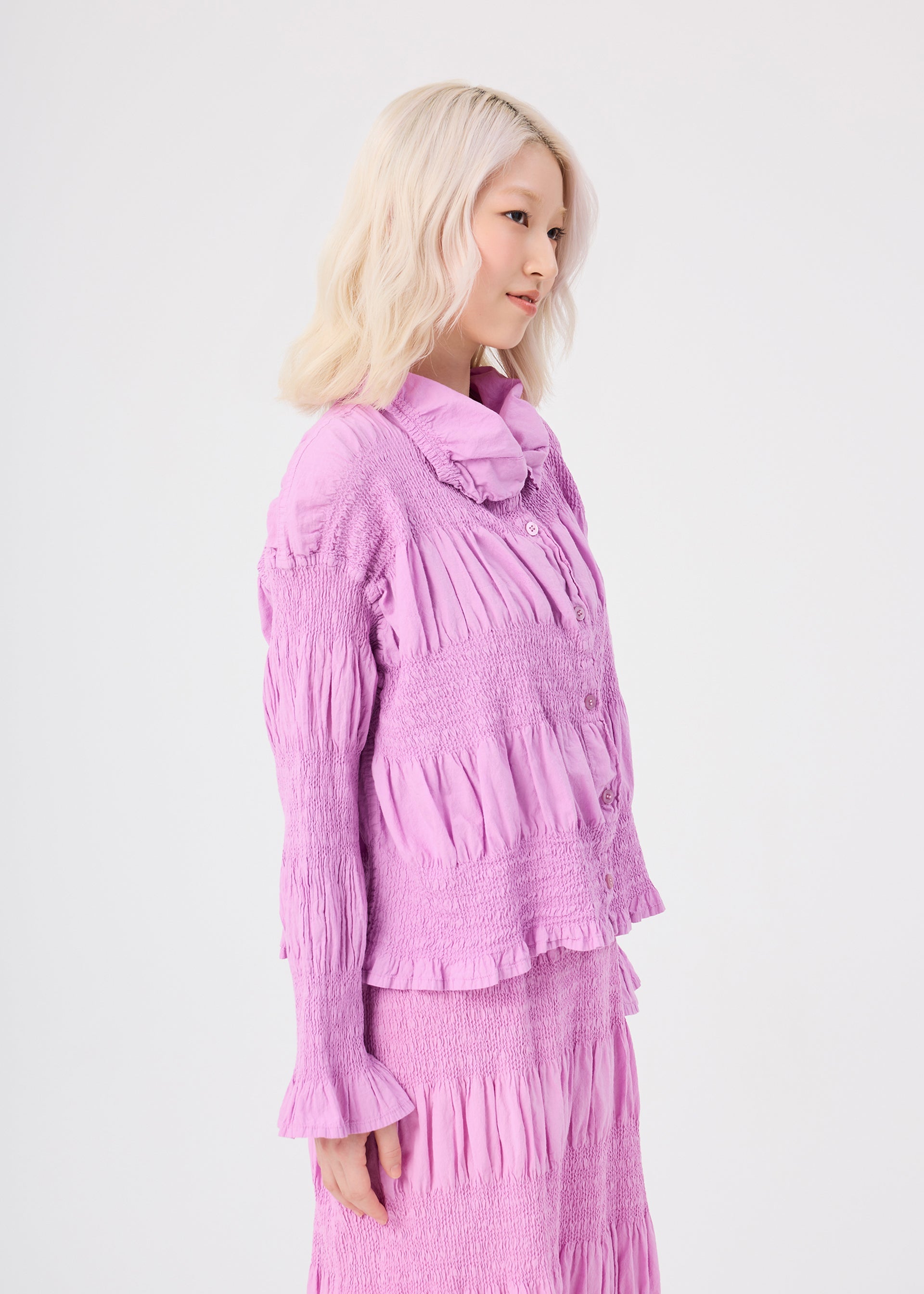 Zoomed view A woman with light blonde hair stands sideways against a plain white background, wearing a textured, long-sleeved lavender blouse and matching skirt, complemented by the me ISSEY MIYAKE SHRINK STRIPE cardigan, distinguished by its striped pattern and unique product-dyeing technique.