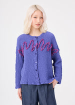 A woman with platinum blonde hair wears the me ISSEY MIYAKE LEAF TULLE CAULIFLOWER, a textured purple cotton cardigan featuring pink ruffle details and tulle tape embroidery, paired with blue pants, standing against a plain white background.
