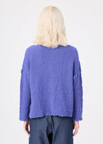 A person with light blonde, wavy hair is shown from behind, wearing a textured, loose-fitting purple LEAF TULLE CAULIFLOWER cardigan by me ISSEY MIYAKE and dark wide-leg pants, standing against a plain white background.