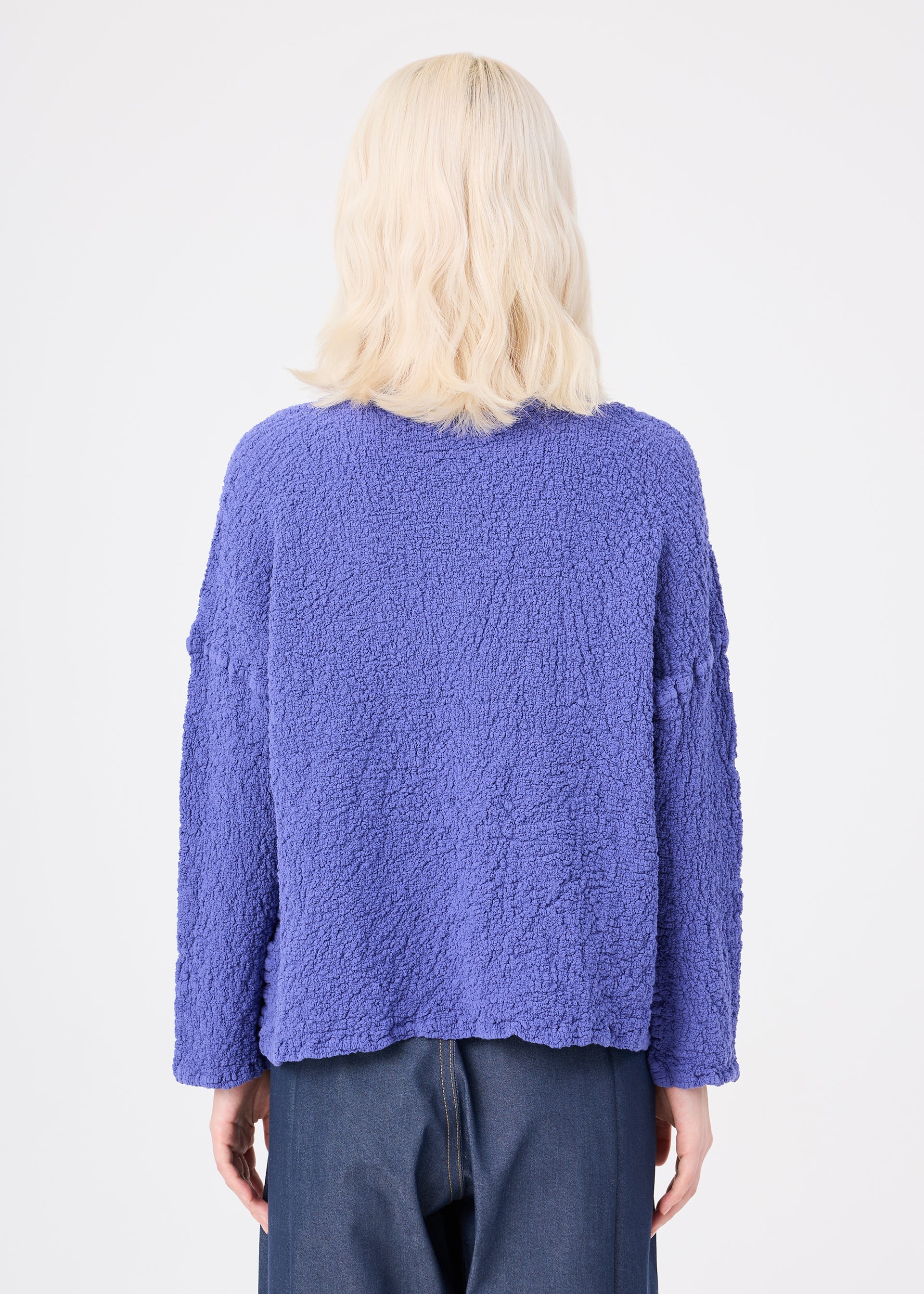 Zoomed view A person with light blonde, wavy hair is shown from behind, wearing a textured, loose-fitting purple LEAF TULLE CAULIFLOWER cardigan by me ISSEY MIYAKE and dark wide-leg pants, standing against a plain white background.