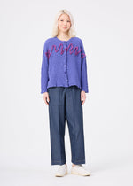 Wearing the LEAF TULLE CAULIFLOWER textured purple cotton cardigan with pink accents and delicate tulle tape embroidery by me ISSEY MIYAKE, a person with light blonde hair pairs it with wide-leg blue trousers and white shoes, standing against a plain white background.