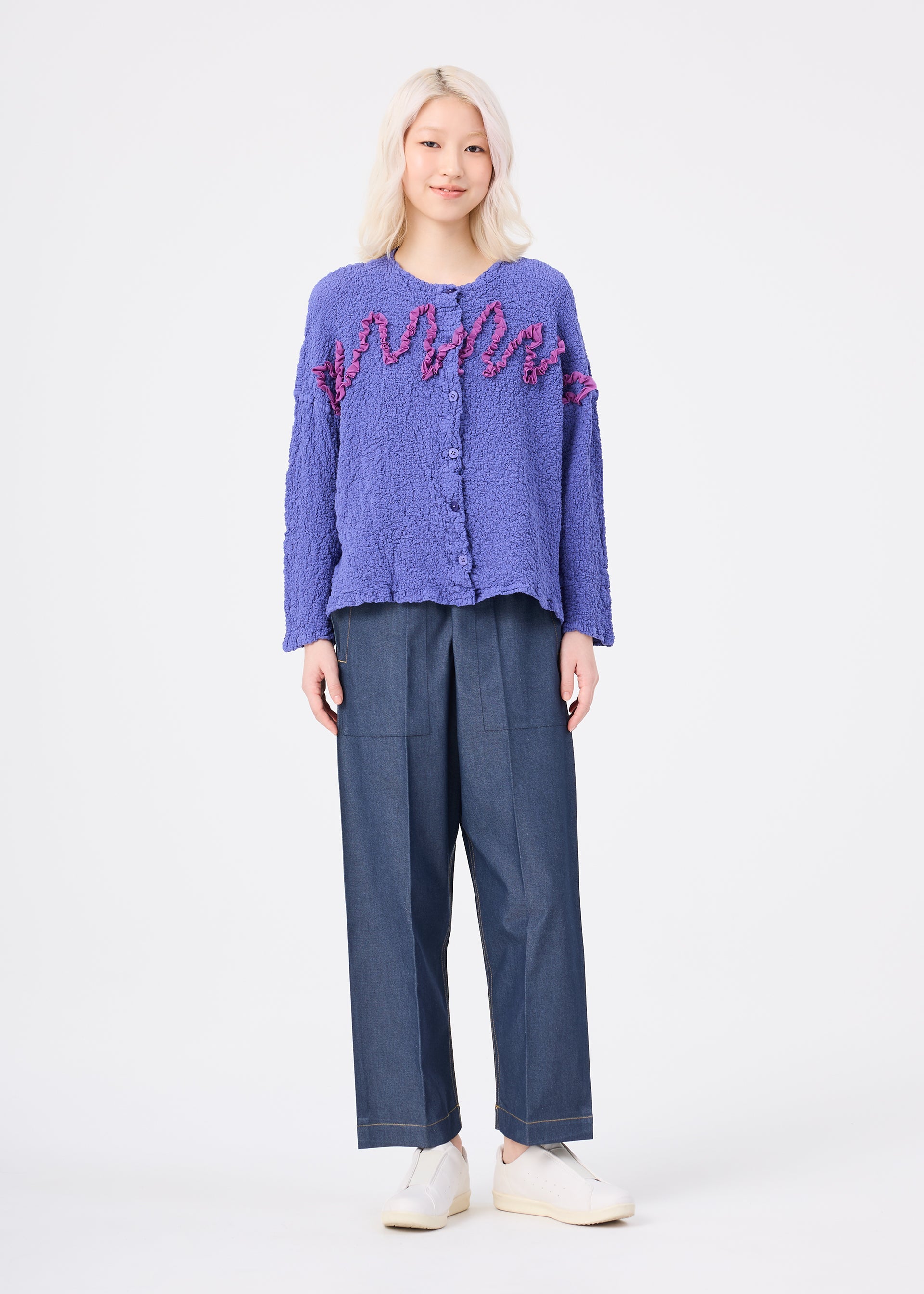 Zoomed view Wearing the LEAF TULLE CAULIFLOWER textured purple cotton cardigan with pink accents and delicate tulle tape embroidery by me ISSEY MIYAKE, a person with light blonde hair pairs it with wide-leg blue trousers and white shoes, standing against a plain white background.