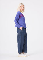 A person with light blonde hair wears the LEAF TULLE CAULIFLOWER textured purple sweater with pink tulle tape embroidery accents from me ISSEY MIYAKE, paired with wide-leg blue pants and white shoes, standing and smiling against a plain white background.