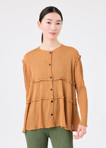 A woman wearing the FLARE SEE-THROUGH CREPE blouse by me ISSEY MIYAKE—a loose, textured brown button-up top crafted from natural plant dyes with ruffle details—paired with green pants stands facing the camera against a plain white background.