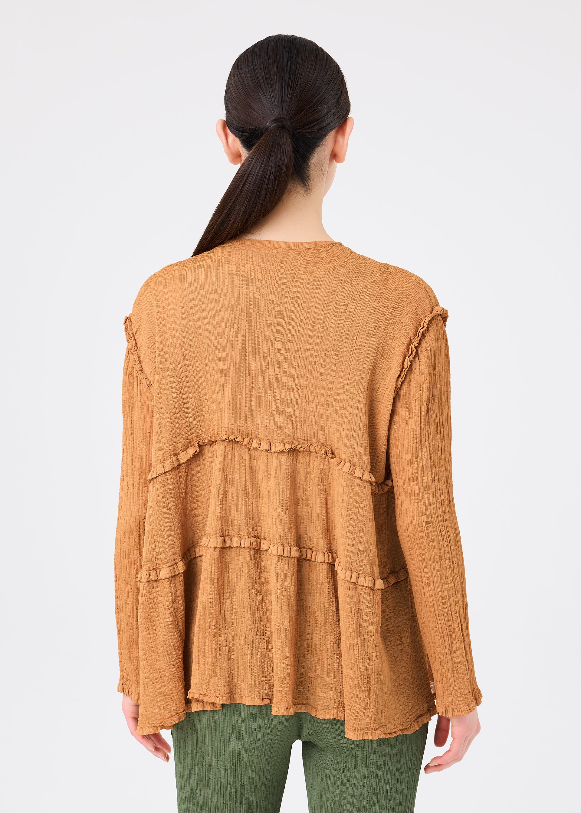 Zoomed view A person with long dark hair in a low ponytail wears the FLARE SEE-THROUGH CREPE top by me ISSEY MIYAKE—a loose, textured brown long-sleeve top crafted from upcycled garments featuring ruffle details—paired with green pants, standing with their back to the camera against a plain white background.