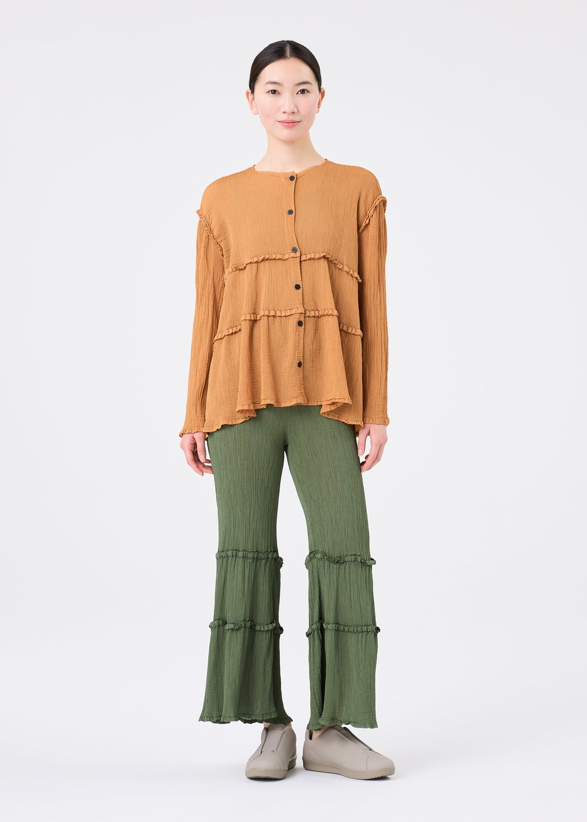 A person stands facing forward against a plain white background wearing the me ISSEY MIYAKE FLARE SEE-THROUGH CREPE, styled in loose mustard tones with long sleeves and natural plant dye, paired with green tiered flared pants and light gray shoes. view 7 of 7