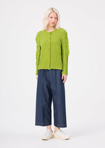 A person with light blonde hair wears the me ISSEY MIYAKE ROUND CAULIFLOWER lime green stretch pleats cardigan, wide-leg dark blue pants, and white slip-on shoes against a plain white background.