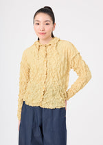 A woman with long dark hair in a ponytail wears the MERINGUE TOP by me ISSEY MIYAKE, a textured yellow polyester top with ruffled edges, paired with loose blue pants against a plain white background.