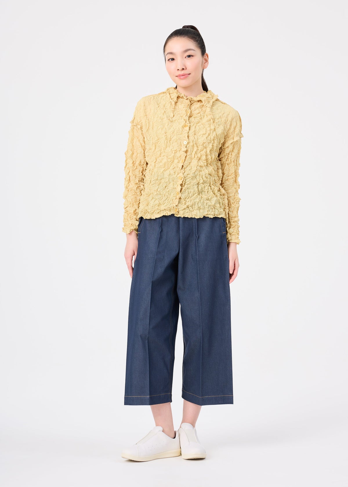 A person stands against a white background wearing the MERINGUE TOP by me ISSEY MIYAKE, paired with wide-leg blue cropped pants and white sneakers, arms relaxed at their sides. view 10 of 10
