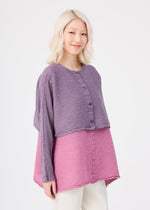 A person with light blonde hair wears the me ISSEY MIYAKE POLYCOTTON CREPE two-tone purple and pink top with front buttons and white pants, standing against a plain white background, smiling gently.