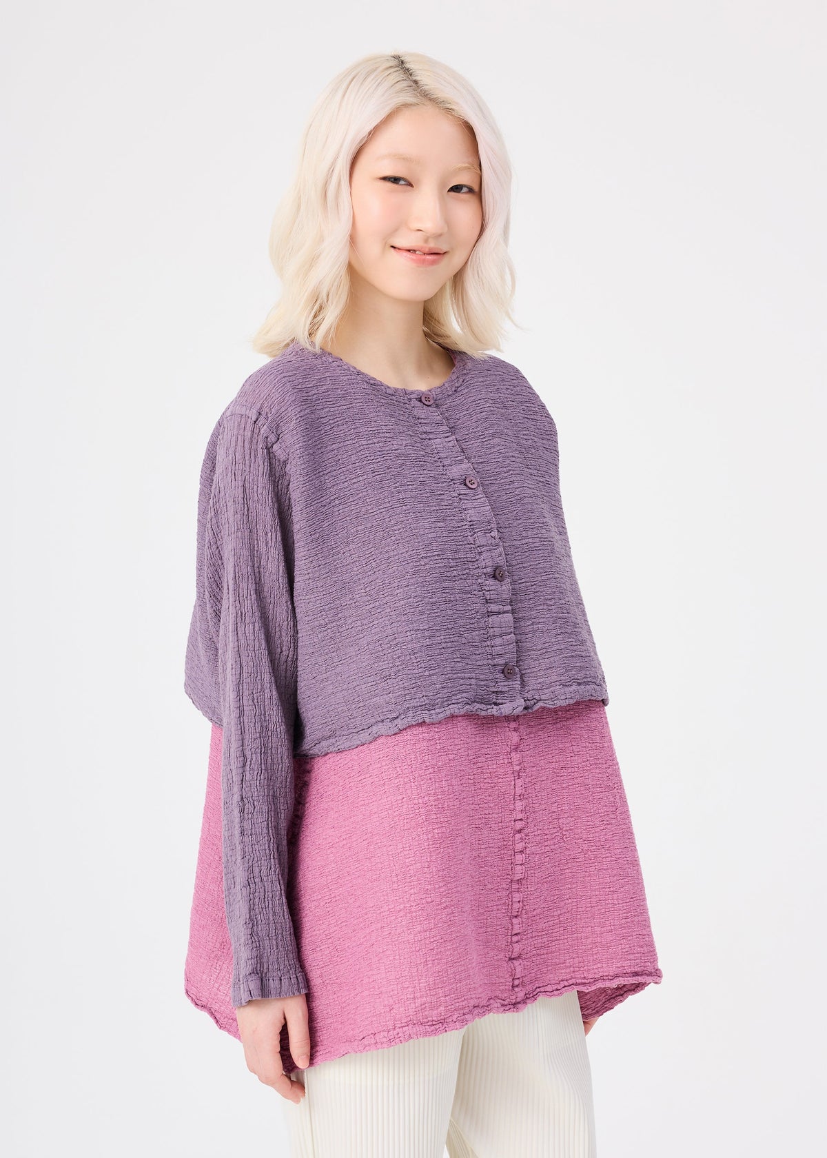 Zoomed view A person with light blonde hair wears the me ISSEY MIYAKE POLYCOTTON CREPE two-tone purple and pink top with front buttons and white pants, standing against a plain white background, smiling gently.
