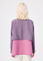 A person with wavy, shoulder-length blonde hair is seen from the back wearing the me ISSEY MIYAKE POLYCOTTON CREPE jacket in purple and pink, paired with light pants, against a plain white background.