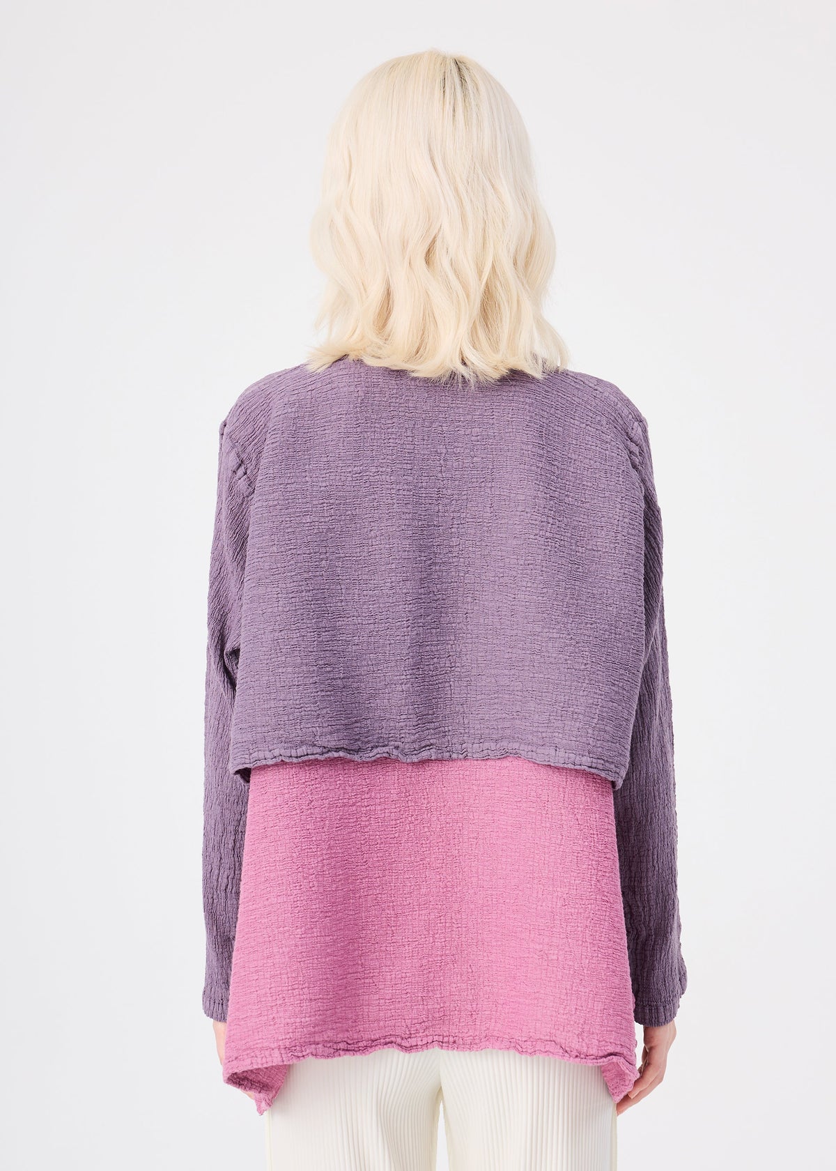 Zoomed view A person with wavy, shoulder-length blonde hair is seen from the back wearing the me ISSEY MIYAKE POLYCOTTON CREPE jacket in purple and pink, paired with light pants, against a plain white background.