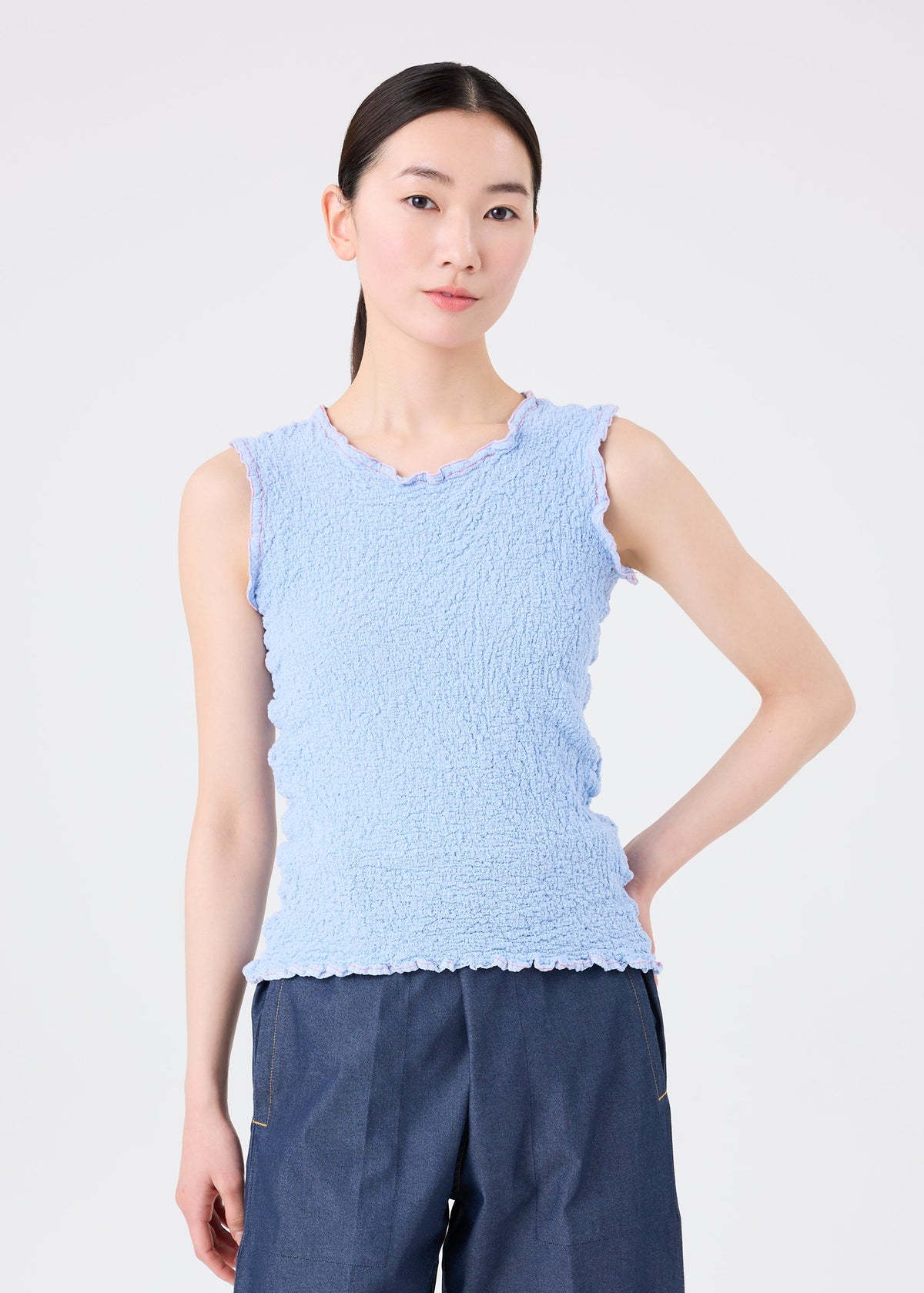 A woman with straight black hair wears the CAULIFLOWER COLOR 1 sleeveless top by me ISSEY MIYAKE in light blue, styled with dark blue pants. She stands against a plain white background, her expression neutral. view 5 of 7