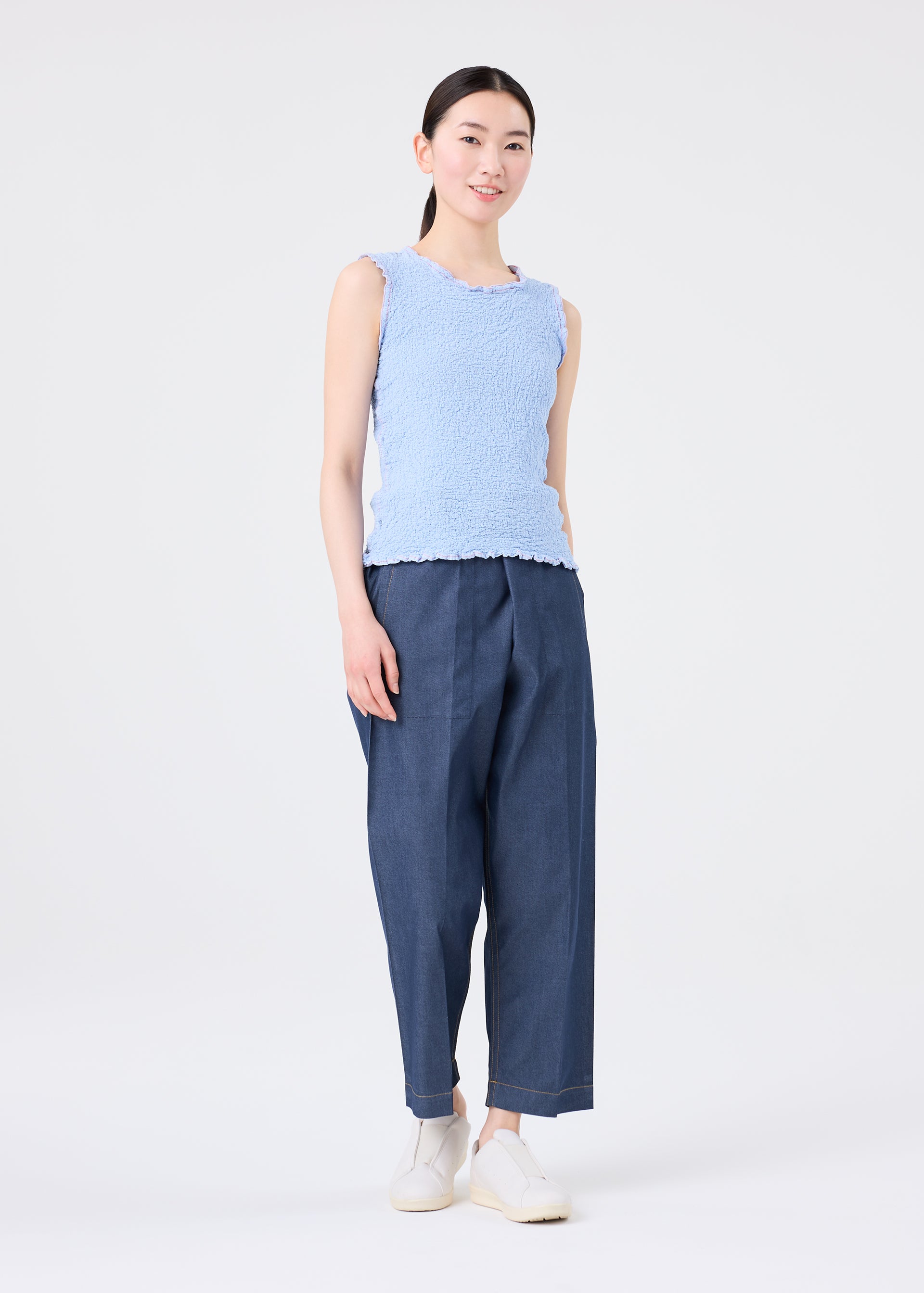 Zoomed view A woman with long dark hair pulled back wears the CAULIFLOWER COLOR 1 sleeveless top by me ISSEY MIYAKE, paired with loose dark blue pants and white slip-on shoes, standing against a plain white background and smiling slightly.