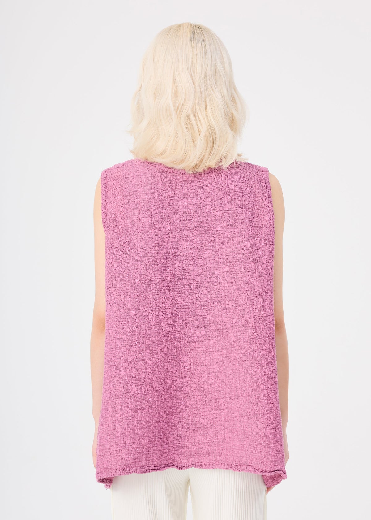 A person with shoulder-length blonde hair is seen from behind wearing the me ISSEY MIYAKE POLYCOTTON CREPE sleeveless pink textured tunic and off-white pants against a plain white background. view 7 of 8