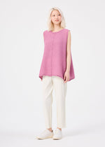 A person with light hair wears the me ISSEY MIYAKE POLYCOTTON CREPE, a loose, sleeveless pink tunic paired with white pants and shoes, standing against a plain white background.