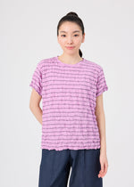 A person stands against a plain background, wearing the STRIPE MERINGUE T-SHIRT by me ISSEY MIYAKE—a light purple, round-neck top with horizontal stripes and a subtle crushed finish that brings dimension to its polyester jersey fabric—paired with dark blue pants. Their long dark hair is pulled back, and they have a neutral expression.