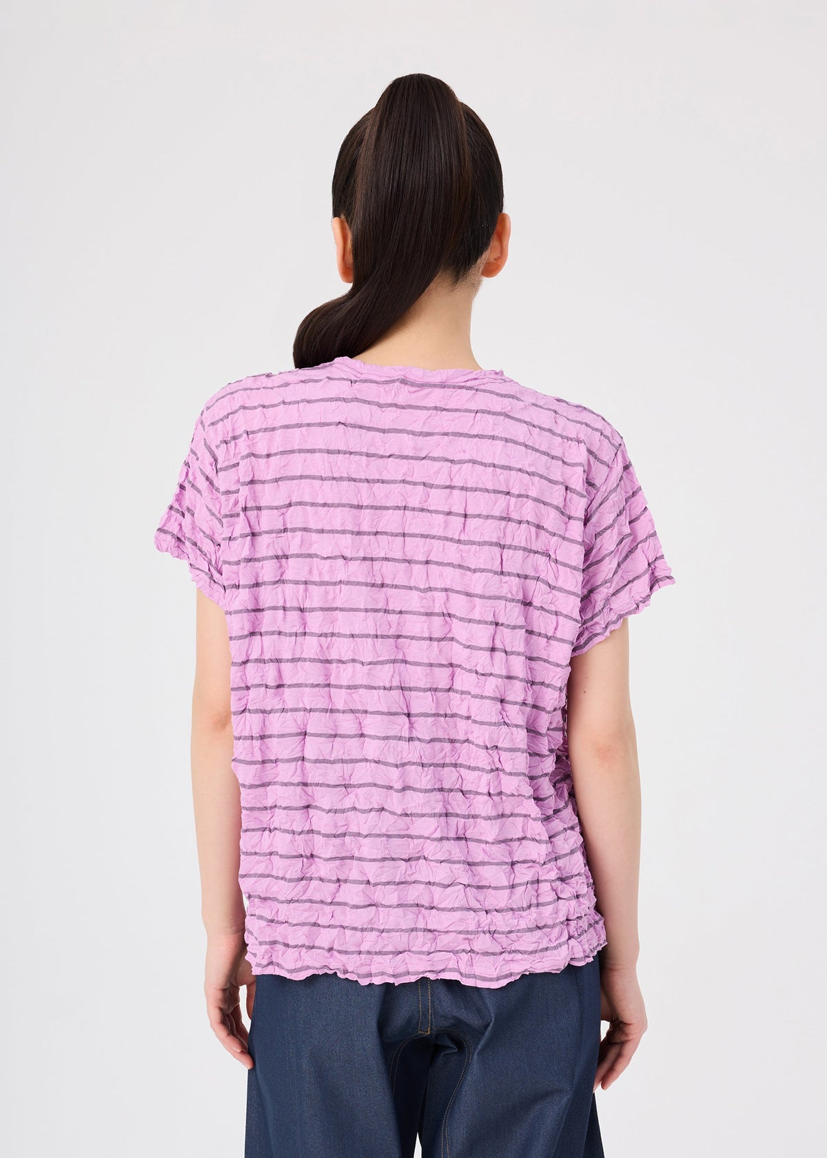 A person with long dark hair in a ponytail is seen from behind, wearing the me ISSEY MIYAKE STRIPE MERINGUE T-SHIRT—a textured, short-sleeve light purple polyester jersey round-neck top—paired with dark pants, standing against a plain white background. view 6 of 7