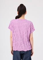 A person with long dark hair in a ponytail is seen from behind, wearing the me ISSEY MIYAKE STRIPE MERINGUE T-SHIRT—a textured, short-sleeve light purple polyester jersey round-neck top—paired with dark pants, standing against a plain white background.