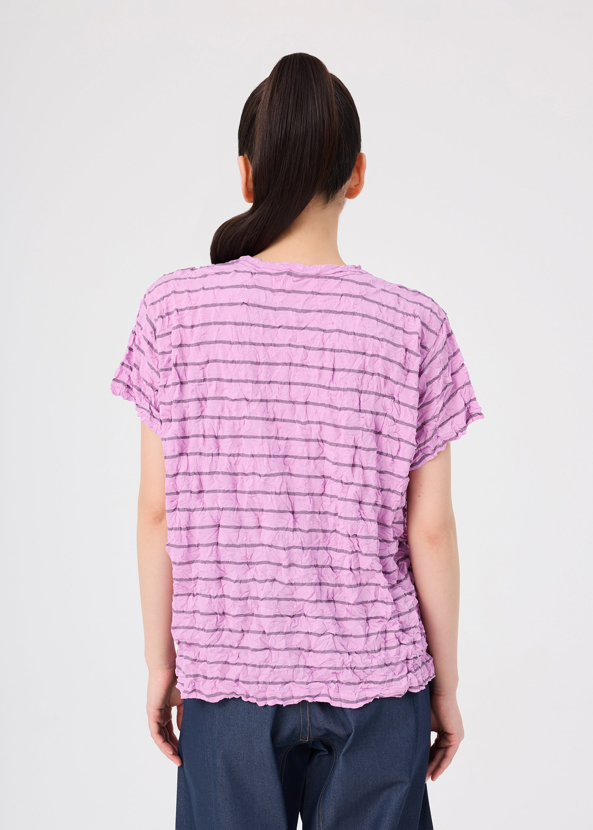 Zoomed view A person with long dark hair in a ponytail is seen from behind, wearing the me ISSEY MIYAKE STRIPE MERINGUE T-SHIRT—a textured, short-sleeve light purple polyester jersey round-neck top—paired with dark pants, standing against a plain white background.