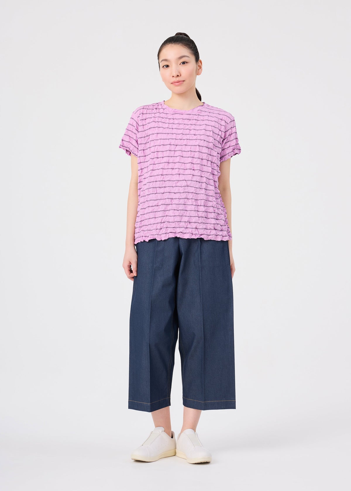 A woman stands facing forward, wearing the STRIPE MERINGUE T-SHIRT by me ISSEY MIYAKE—a ruffled pink round-neck top with a subtle crushed finish—paired with wide-leg blue denim pants and white slip-on shoes, against a plain white background. view 7 of 7