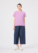 A woman stands facing forward, wearing the STRIPE MERINGUE T-SHIRT by me ISSEY MIYAKE—a ruffled pink round-neck top with a subtle crushed finish—paired with wide-leg blue denim pants and white slip-on shoes, against a plain white background.