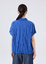 A person with long dark hair in a low ponytail is shown from the back against a plain light background, wearing the STRIPE MERINGUE T-SHIRT by me ISSEY MIYAKE—a textured blue polyester jersey shirt—paired with dark pants.