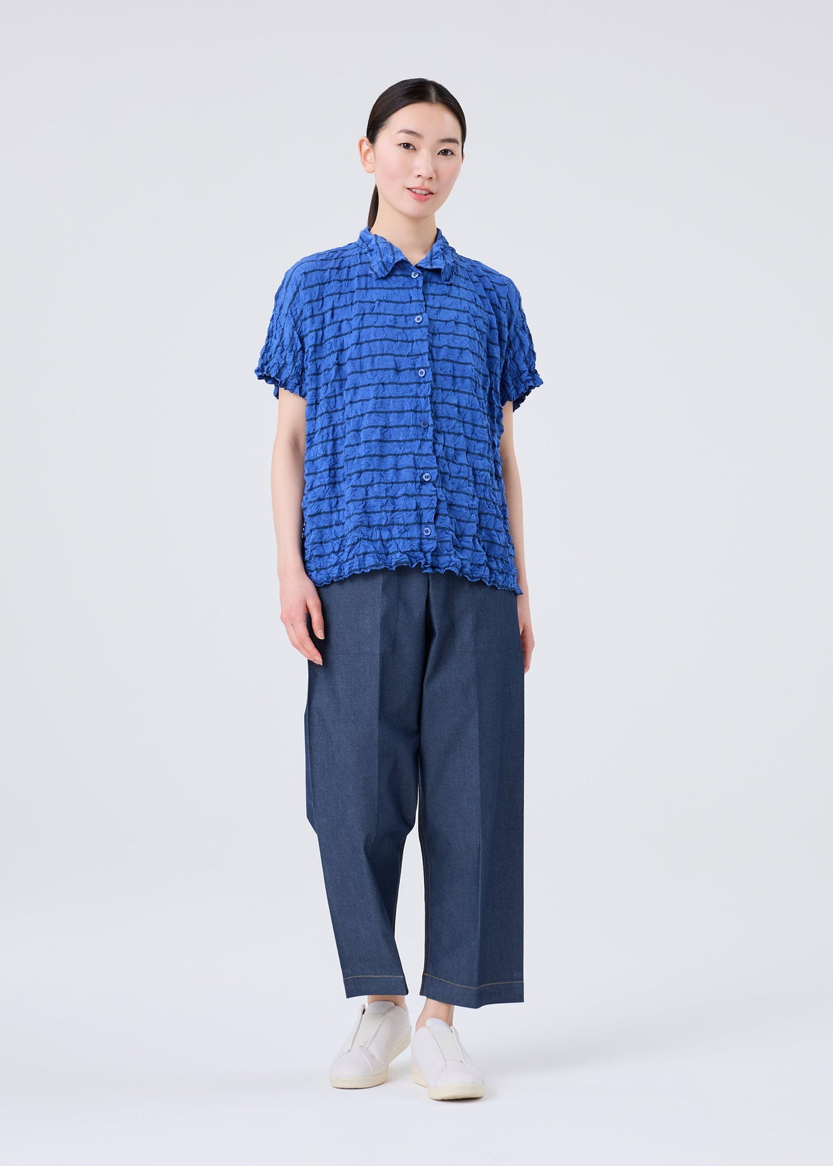 A person stands facing forward against a plain light background, wearing the STRIPE MERINGUE T-SHIRT by me ISSEY MIYAKE—a textured blue short-sleeve shirt with a striped pattern—paired with loose blue pants and white shoes. view 7 of 7