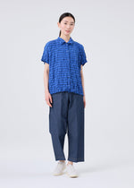 A person stands facing forward against a plain light background, wearing the STRIPE MERINGUE T-SHIRT by me ISSEY MIYAKE—a textured blue short-sleeve shirt with a striped pattern—paired with loose blue pants and white shoes.