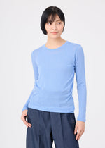 A woman with short black hair wears the me ISSEY MIYAKE WOOL MIX JERSEY in light blue, paired with dark blue pants, standing against a white background and gazing slightly left with a neutral expression.