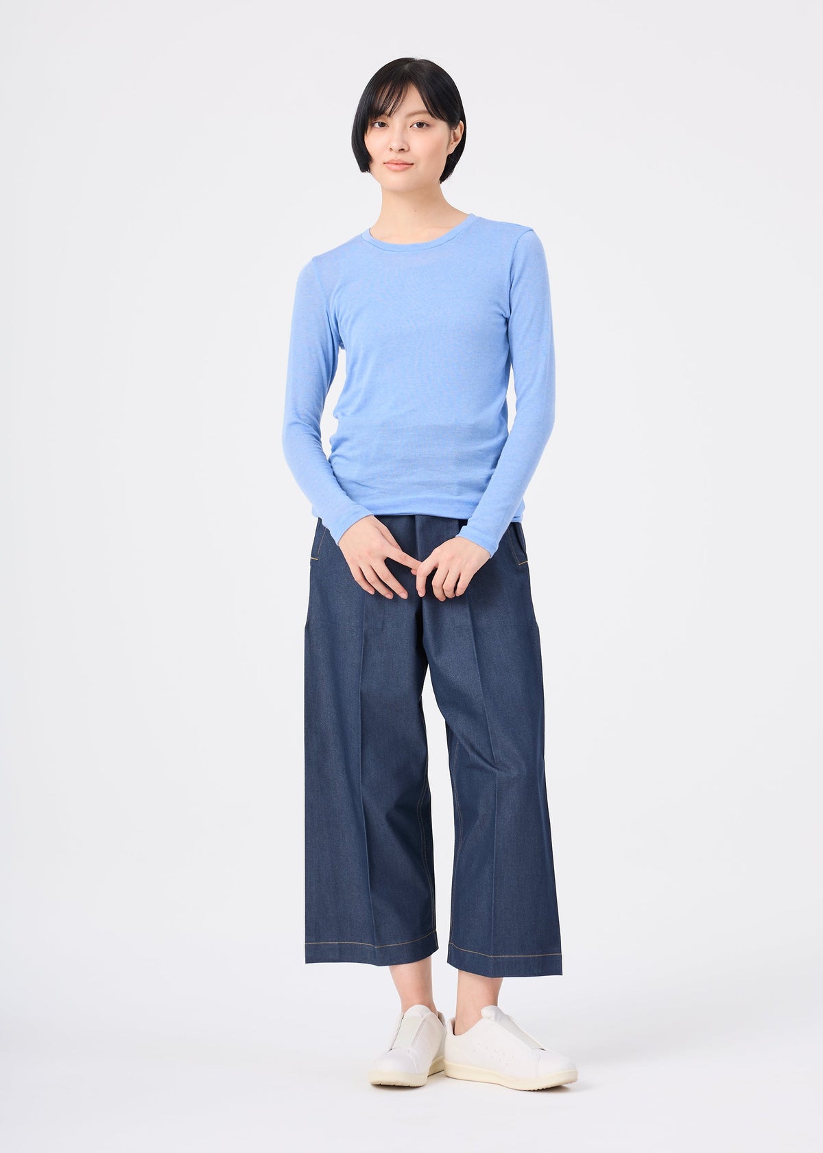 Zoomed view A person with short black hair wears the me ISSEY MIYAKE WOOL MIX JERSEY in light blue, wide-leg dark denim pants, and white slip-on shoes, standing against a plain white background.