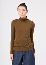 A person stands against a plain white background wearing the me ISSEY MIYAKE WOOL MIX JERSEY, a brown long-sleeved high-neck top, and blue pants, facing forward with a slight smile and hair tied back.