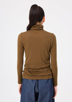 A person with long dark hair in a ponytail wears the me ISSEY MIYAKE WOOL MIX JERSEY top—brown, long-sleeved, and high-necked—with blue trousers, standing with their back to the camera against a white background.